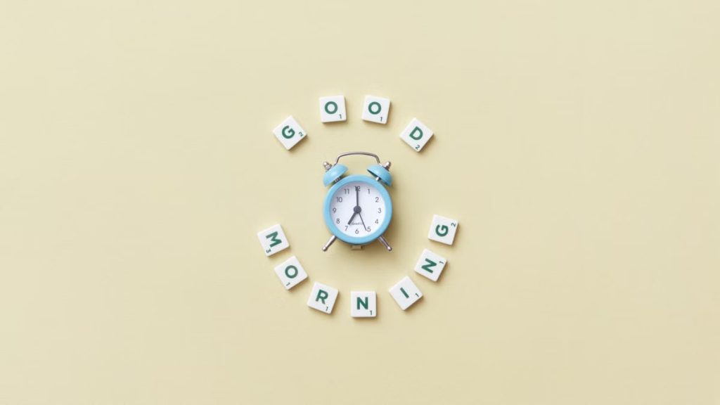 Aesthetic flat lay with a blue alarm clock surrounded by scrabble tiles spelling 'Good Morning' on a pastel background.