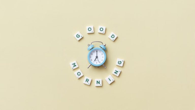 Aesthetic flat lay with a blue alarm clock surrounded by scrabble tiles spelling 'Good Morning' on a pastel background.